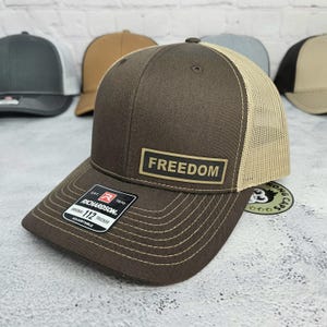 May include: Brown trucker hat with a tan mesh back and a curved brim. The hat features the word "FREEDOM" on a rectangular patch. The Richardson 112 Trucker hat is adjustable.