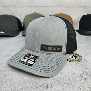 May include: A gray trucker hat with a black mesh back and a curved brim. The hat features a rectangular patch with the word "FREEDOM" and a Richardson 112 Trucker label. Other hats are visible in the background.