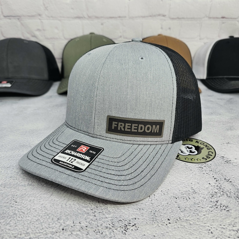 May include: A gray trucker hat with a black mesh back and a curved brim. The hat features a rectangular patch with the word "FREEDOM" and a Richardson 112 Trucker label. Other hats are visible in the background.