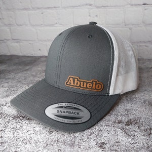 May include: A gray snapback hat with a white mesh back and a curved brim. The hat features the word "Abuelo" in a brown, leather-like patch on the front. The hat has a snap closure and a "Yupoong" label.