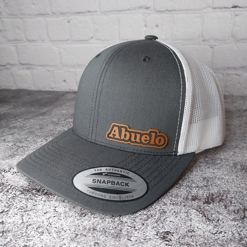 May include: A gray snapback hat with a white mesh back and a curved brim. The hat features the word "Abuelo" in a brown, leather-like patch on the front. The hat has a snap closure and a "Yupoong" label.