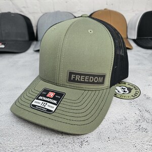 May include: Olive green trucker hat with a black mesh back and a curved brim. The hat features a rectangular patch with the word "FREEDOM" and a Richardson patch. Several other hats are visible in the background.