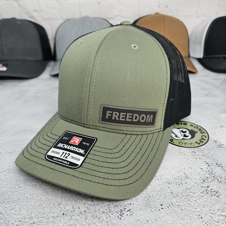 May include: Olive green trucker hat with a black mesh back and a curved brim. The hat features a rectangular patch with the word "FREEDOM" and a Richardson patch. Several other hats are visible in the background.