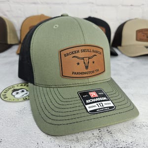 May include: A green and black trucker hat with a brown leather patch that reads "BROKEN SKULL RANCH FARMINGTON TN". The hat has a curved brim and an adjustable strap. The brand is Richardson.