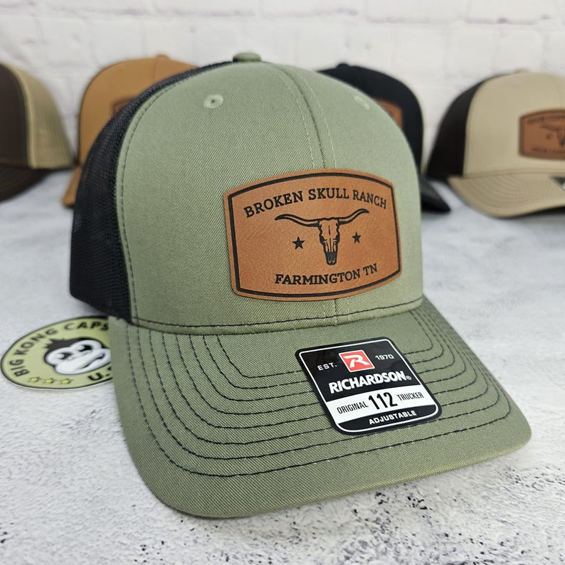 May include: A green and black trucker hat with a brown leather patch that reads "BROKEN SKULL RANCH FARMINGTON TN". The hat has a curved brim and an adjustable strap. The brand is Richardson.