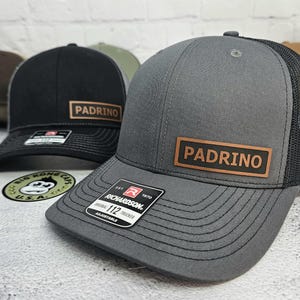 May include: Gray and black trucker hats with the word "PADRINO" on a brown leather patch. The hats have a mesh back and a curved brim. The Richardson 112 trucker hat is adjustable.