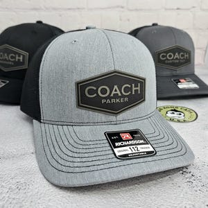 May include: A gray trucker hat with a black mesh back and a curved brim. The hat features a black patch with the word "COACH" and "PARKER" in white. The hat has a "RICHARDSON 112 TRUCKER" tag.