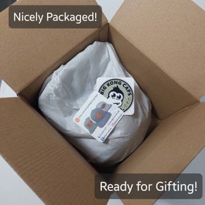 May include: A brown cardboard box containing a wrapped item, with the text "Nicely Packaged!" and "Ready for Gifting!". The item is wrapped in white tissue paper and includes a sticker with a gorilla logo and the words "Big Kong Caps".