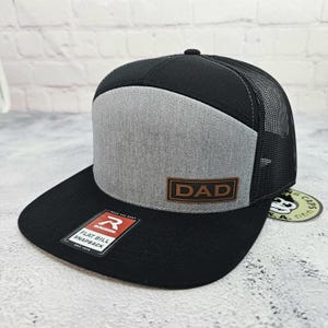 May include: Black and gray flat bill snapback hat with a mesh back. The front panel is gray with a brown leather patch that reads "DAD". The brim is black, and the hat has a Richardson tag.