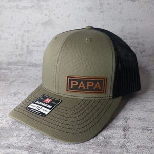 May include: Olive green and black trucker hat with the word "PAPA" on a brown leather patch. The hat has a curved brim and a black mesh back. The Richardson 112 adjustable hat is a classic style.