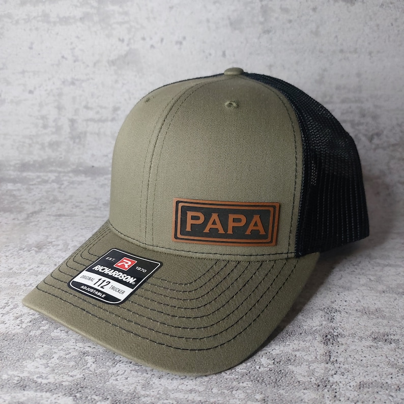 May include: Olive green and black trucker hat with the word "PAPA" on a brown leather patch. The hat has a curved brim and a black mesh back. The Richardson 112 adjustable hat is a classic style.
