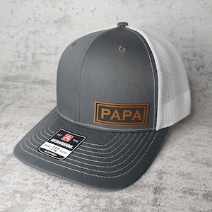 May include: A gray and white trucker hat with the word "PAPA" on a brown leather patch. The hat has a curved brim and a mesh back. The Richardson brand tag is visible.