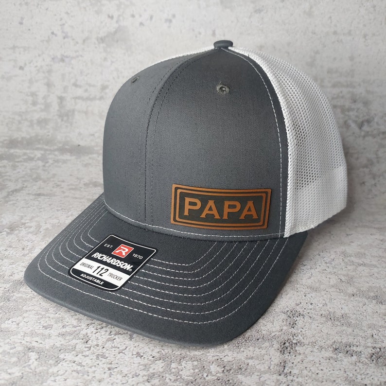 May include: A gray and white trucker hat with the word "PAPA" on a brown leather patch. The hat has a curved brim and a mesh back. The Richardson brand tag is visible.