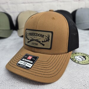 May include: A brown trucker hat with a black mesh back. The front panel features a rectangular patch with the word "FREEDOM" above crossed rifles and antlers, with "1776" below. The hat has a curved brim and an adjustable strap.