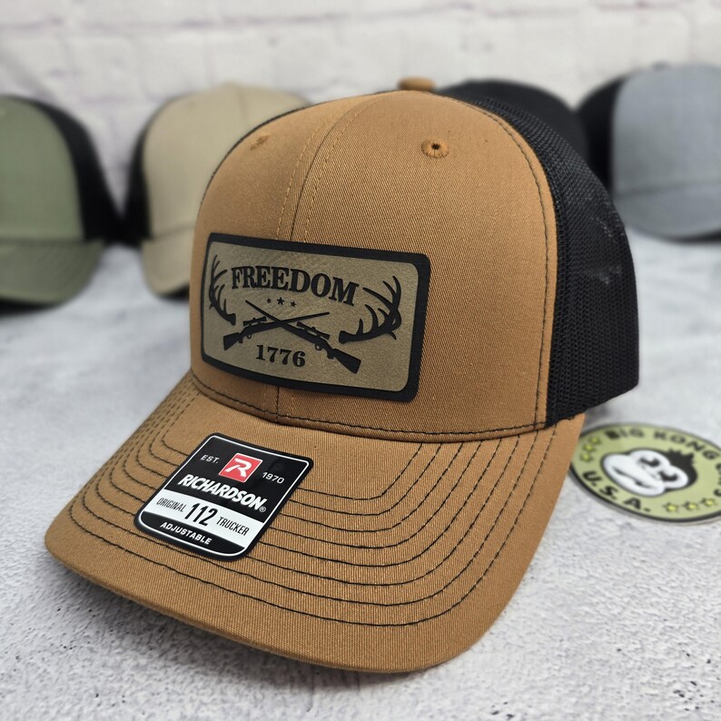 May include: A brown trucker hat with a black mesh back. The front panel features a rectangular patch with the word "FREEDOM" above crossed rifles and antlers, with "1776" below. The hat has a curved brim and an adjustable strap.