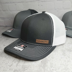 May include: Black and white trucker hat with a leather patch that reads "GRANDPA". The hat has a curved brim and a mesh back. The hat is adjustable and has a Richardson 112 trucker tag.