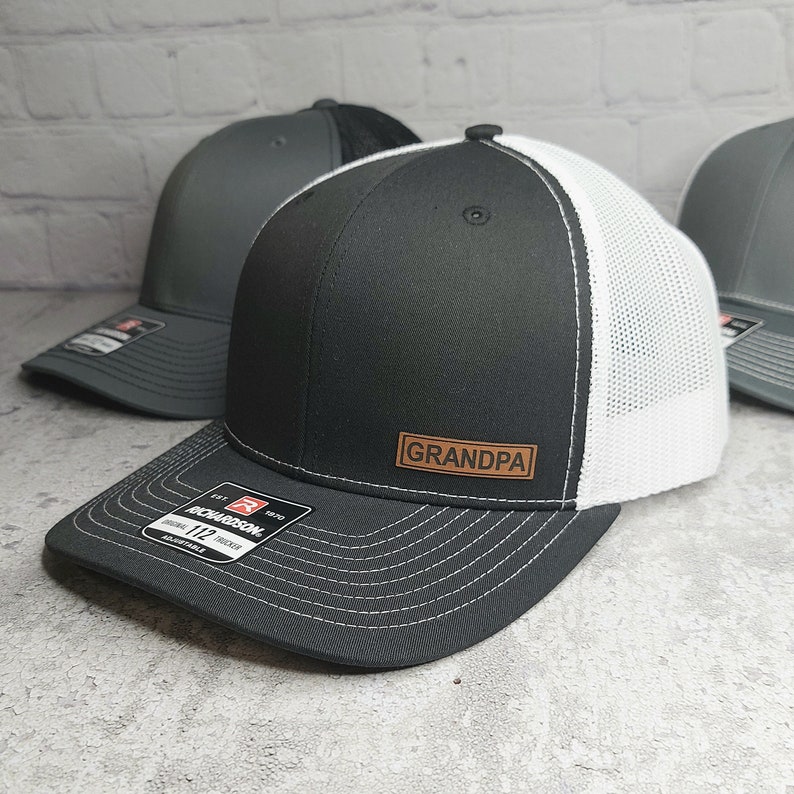 May include: Black and white trucker hat with a leather patch that reads "GRANDPA". The hat has a curved brim and a mesh back. The hat is adjustable and has a Richardson 112 trucker tag.