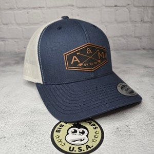 May include: A navy blue and white trucker hat with a brown leather patch that says "A & M 07.22.25" with two arrows crossing each other.
