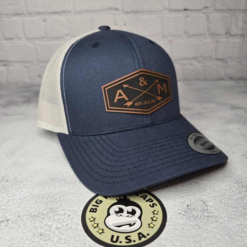 May include: A navy blue and white trucker hat with a brown leather patch that says "A & M 07.22.25" with two arrows crossing each other.