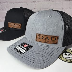 May include: Two trucker hats, one black and one gray, with mesh backs. Each hat features a brown leather patch with the word "DAD" and an established year. The black hat says "EST. 2026" and the gray hat says "EST. 2027".