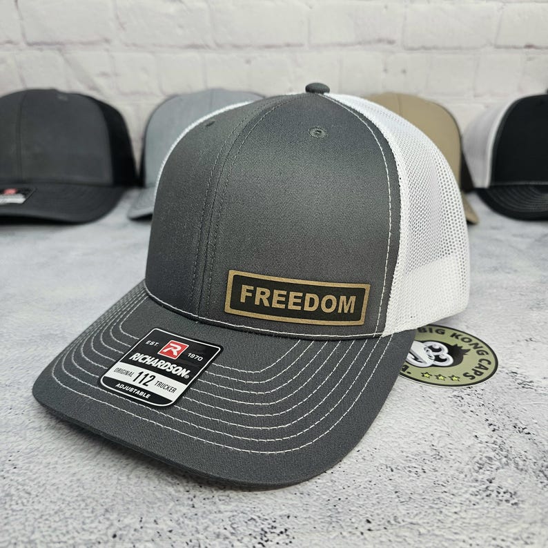May include: A gray Richardson 112 trucker hat with a white mesh back and a curved brim. The hat features the word "FREEDOM" on a rectangular patch. The hat is adjustable.