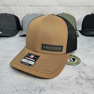 May include: A tan trucker hat with a black mesh back and a curved brim. The hat features a rectangular patch with the word "FREEDOM" and a Richardson 112 Trucker label. Several other hats are visible in the background.