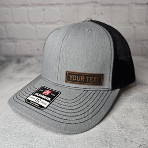 以下が含まれることがあります： グレーとブラックのトラッカーハット。ブラウンの革のパッチには「YOUR TEXT」と書いてあります。ハットはブラックメッシュバック、グレーフロント、ブラックステッチのつば付き。ハットは調節可能で、リチャードソン112のラベルが付いています。