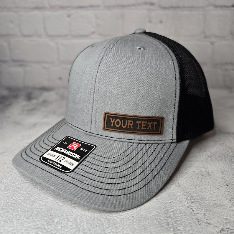 以下が含まれることがあります： グレーとブラックのトラッカーハット。ブラウンの革のパッチには「YOUR TEXT」と書いてあります。ハットはブラックメッシュバック、グレーフロント、ブラックステッチのつば付き。ハットは調節可能で、リチャードソン112のラベルが付いています。