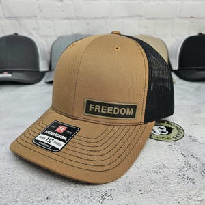 May include: A brown trucker hat with a black mesh back. The hat features the word "FREEDOM" on a rectangular patch. The Richardson brand label is visible on the front. Other hats are in the background.