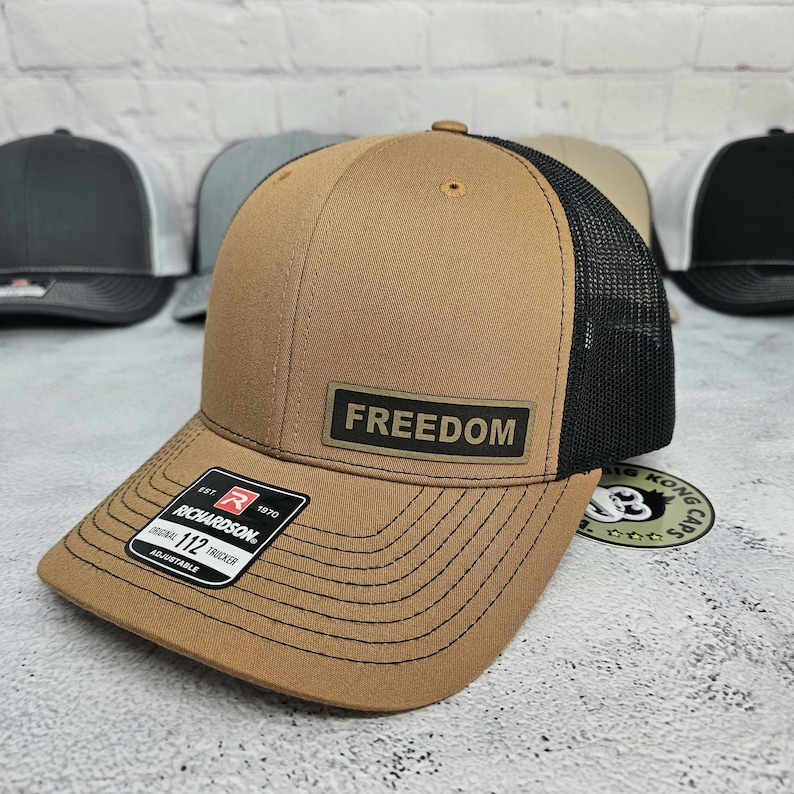 May include: A brown trucker hat with a black mesh back. The hat features the word "FREEDOM" on a rectangular patch. The Richardson brand label is visible on the front. Other hats are in the background.