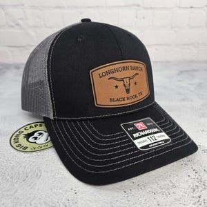 May include: Black trucker hat with a gray mesh back and a brown leather patch. The patch reads "LONGHORN RANCH BLACK ROCK, TX" with a steer skull graphic. Features the Richardson 112 Trucker label.