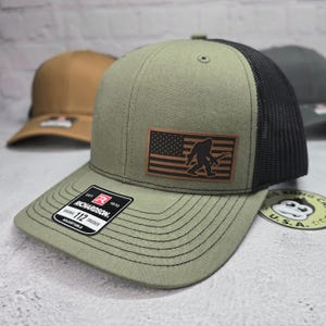 May include: A green and black trucker hat with a brown leather patch featuring an American flag with a silhouette of Bigfoot holding a rifle. The hat has a black mesh back and a green front with the text "Richardson 112 Trucker Adjustable" on the front.