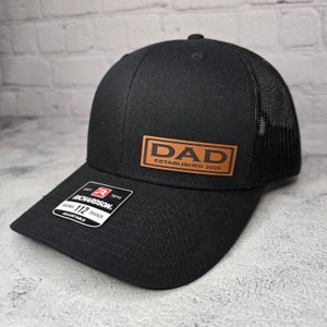 Op de afbeelding: Zwarte truckercap met een bruin leren patch waarop "DAD ESTABLISHED 2025" staat.