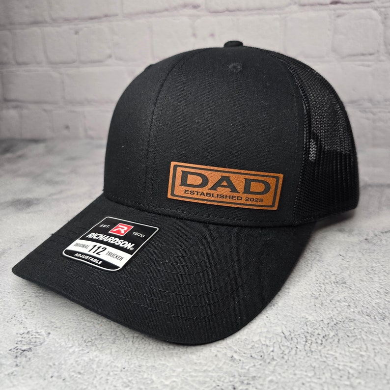 Op de afbeelding: Zwarte truckercap met een bruin leren patch waarop "DAD ESTABLISHED 2025" staat.
