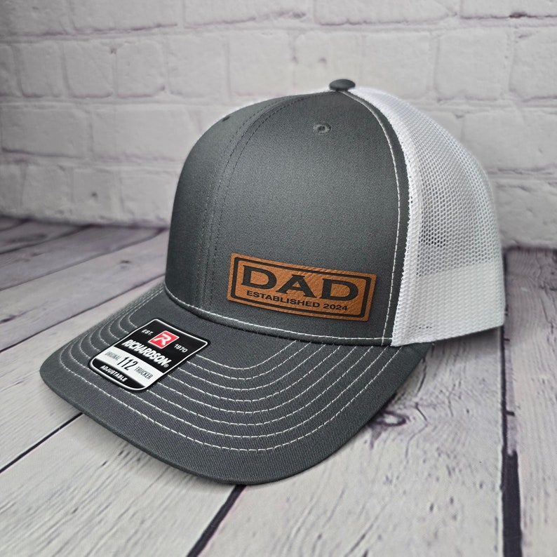 Op de afbeelding: Een grijze en witte truckercap met een bruin leren patch waarop "DAD ESTABLISHED 2024" staat.