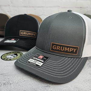 May include: Two trucker hats, one black and one gray, with the word "GRUMPY" on a brown patch. The gray hat has a white mesh back. Both hats have a curved brim and adjustable fit.
