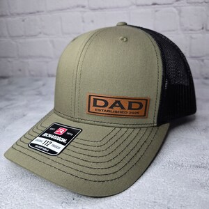 Op de afbeelding: Een groene en zwarte trucker-pet met een bruin leren patch waarop "DAD ESTABLISHED 2025" staat.