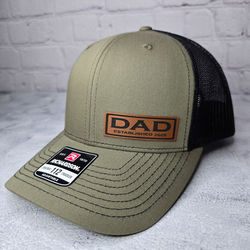 Op de afbeelding: Een groene en zwarte trucker-pet met een bruin leren patch waarop "DAD ESTABLISHED 2025" staat.