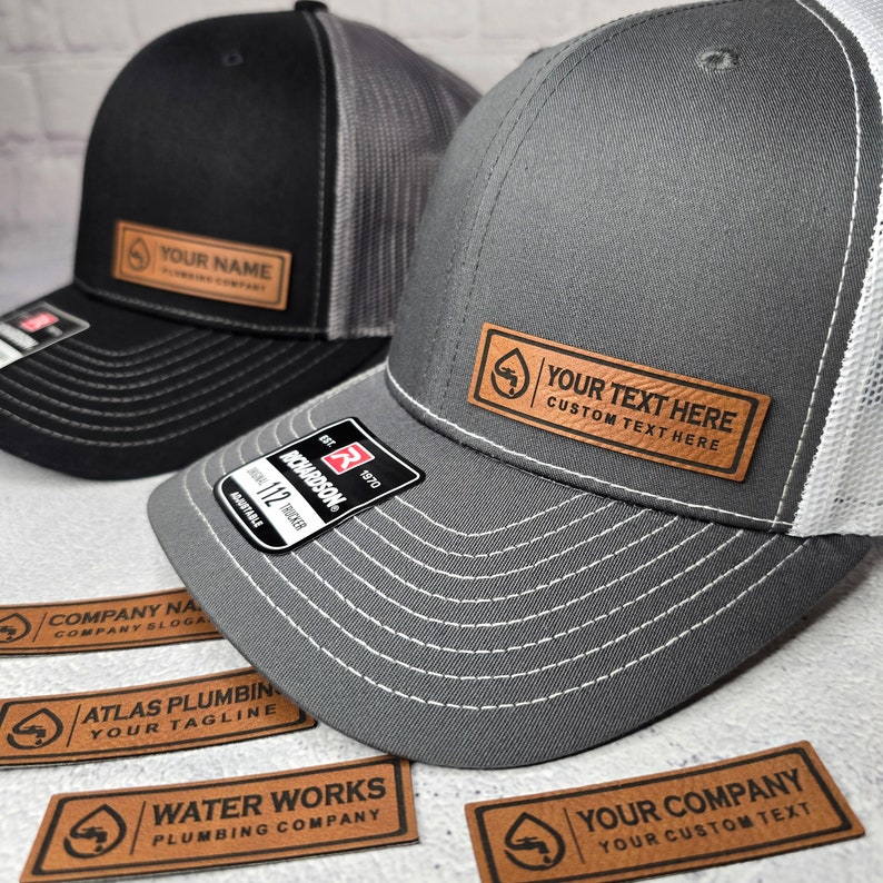 May include: Two trucker hats, one black and one gray, with white mesh backs. Each hat has a leather patch with custom text. The gray hat displays "YOUR TEXT HERE CUSTOM TEXT HERE". Several leather patches with company names and logos are also visible.