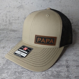 May include: A tan and black trucker hat with the word "PAPA" on a brown leather patch. The hat has a curved brim and a mesh back. The Richardson 112 adjustable hat has a black and white tag.