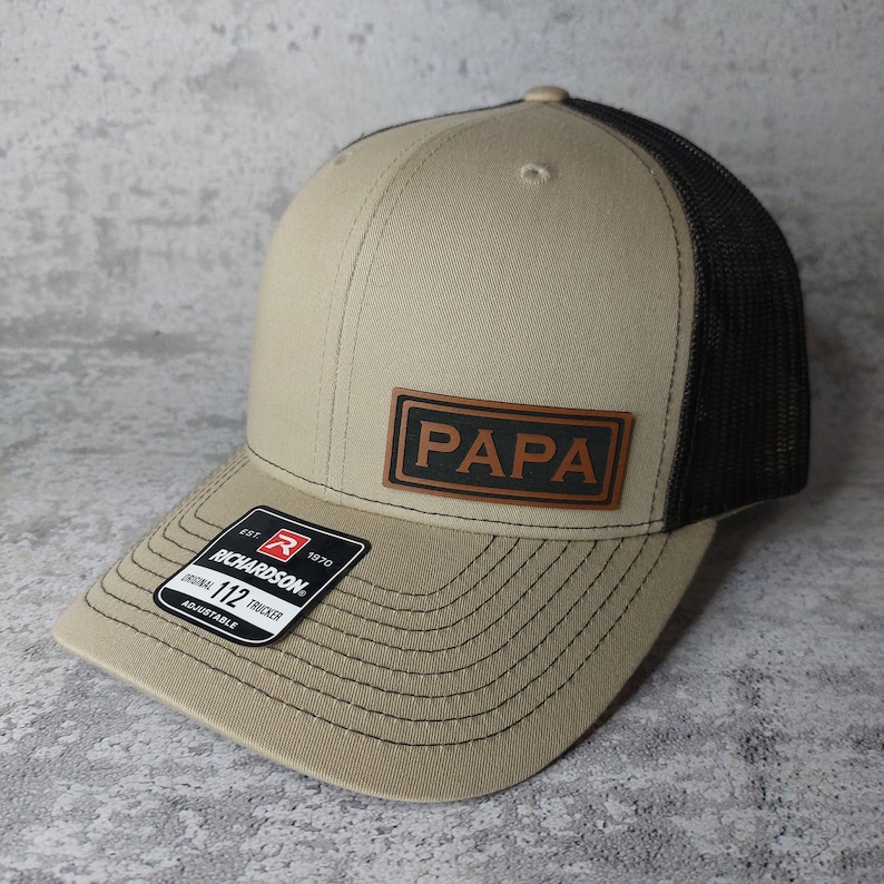 May include: A tan and black trucker hat with the word "PAPA" on a brown leather patch. The hat has a curved brim and a mesh back. The Richardson 112 adjustable hat has a black and white tag.