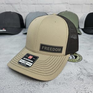 May include: A tan and black trucker hat with the word "FREEDOM" on a rectangular patch. The hat has a mesh back and a curved brim. The Richardson brand label is visible. Other hats are in the background.