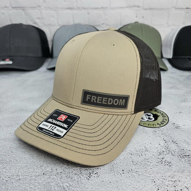 May include: A tan and black trucker hat with the word "FREEDOM" on a rectangular patch. The hat has a mesh back and a curved brim. The Richardson brand label is visible. Other hats are in the background.