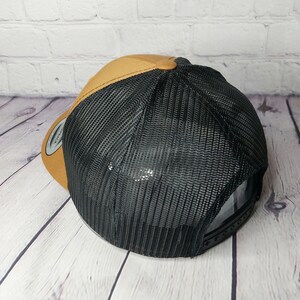 May include: A trucker hat with a brown front panel and a black mesh back. The hat has a curved brim and a snapback closure. The hat is made of a durable material and is designed for comfort and style.