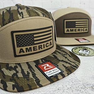 May include: Two flat bill snapback hats. One hat features a camouflage pattern with a tan patch displaying the American flag and the word "AMERICA". The other hat is tan and brown with the same flag design. Both hats have a Richardson tag.