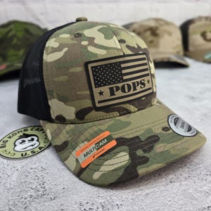 May include: Camouflage baseball cap with a black mesh back and a curved brim. The cap features a rectangular patch with an American flag design and the word "POPS". The cap has a "MULTICAM" sticker.
