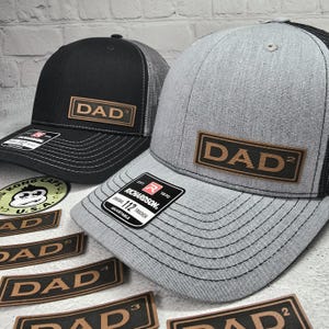 May include: Two trucker hats with a brown leather patch that says "DAD" on them. The hats are one black and one gray. There are several brown leather patches with the word "DAD" on them in the background.