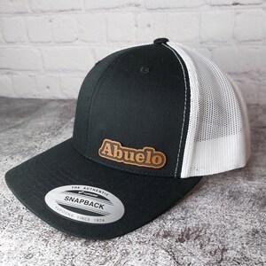May include: Black and white snapback hat with the word "Abuelo" in brown lettering on a brown patch. The hat has a mesh back and a curved brim. The hat also has a "Snapback" tag.