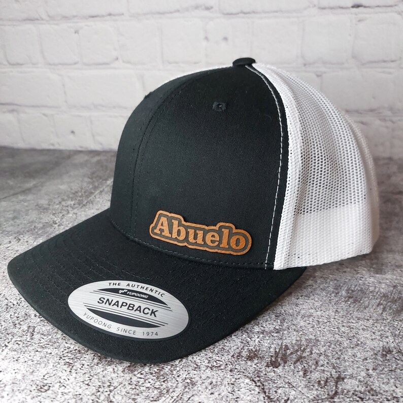 May include: Black and white snapback hat with the word "Abuelo" in brown lettering on a brown patch. The hat has a mesh back and a curved brim. The hat also has a "Snapback" tag.