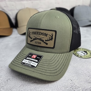 May include: Olive green trucker hat with a black mesh back. The hat features a leather patch with the word "FREEDOM" above "1776" and crossed rifles and antlers. The hat has a curved brim and an adjustable strap.